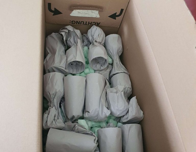 Karton mit mehreren in grauer Packseide eingewickelten Gegenständen und grünem Füllmaterial zum Schutz vor Bruch und Schäden.